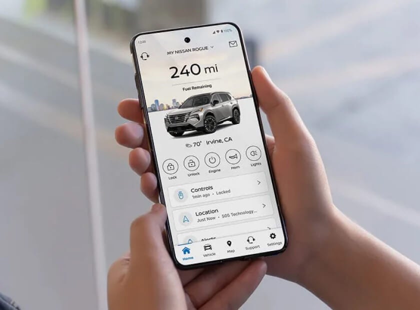 Hands hold a smartphone displaying the MyNISSAN app for a Nissan Rogue, with fuel range, controls and location.