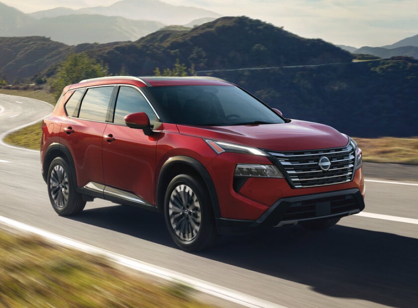 2026 Nissan Rogue Platinum in Scarlet Ember driving on a winding mountain road under a bright sunny sky.